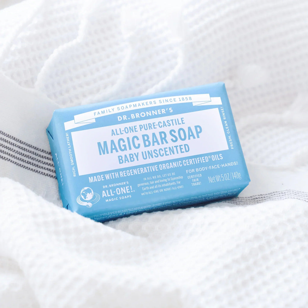 A bar of Dr. Bronners Magic Soap, Baby Unscented, rests on textured white fabric. The packaging is light blue with Organic Ingredients, Certified Fair Trade, Vegan. The setting conveys cleanliness and purity [5 oz]
