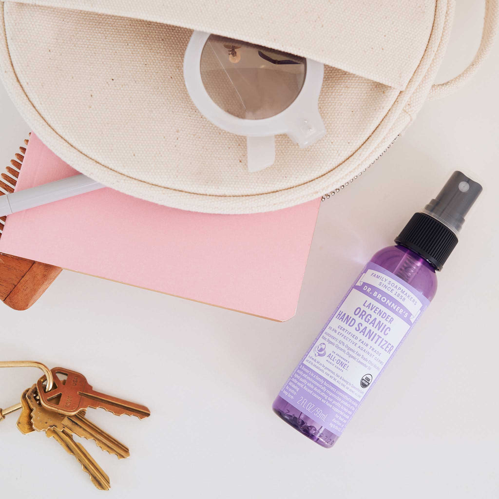 A spray bottle of Dr. Bronners lavender organic hand sanitizer lies beside notebooks, a pouch, and keys on a white surface, highlighting eco friendly daily essentials in a tidy arrangement