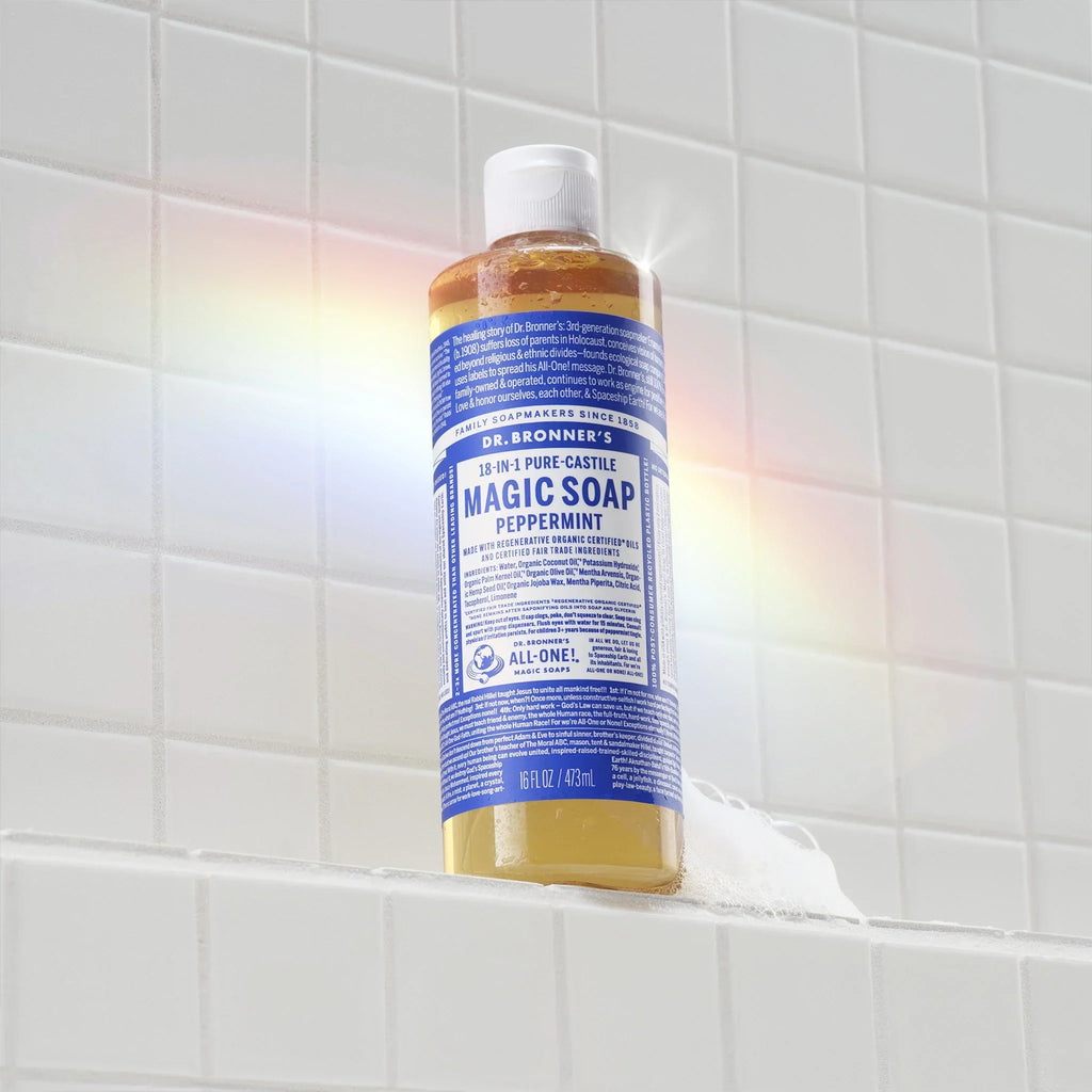 A bottle of Dr. Bronners 18 in 1 Pure Castile Magic Soap in peppermint scent sits on a white tile surface with a subtle rainbow reflection. Text reads Dr. Bronners Magic Soap [16 oz]