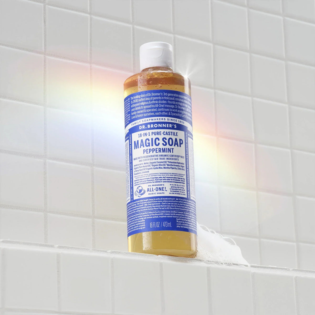 A bottle of Dr. Bronners 18 in 1 Pure Castile Magic Soap in peppermint scent sits on a white tile surface with a subtle rainbow reflection. Text reads Dr. Bronners Magic Soap [16 oz]