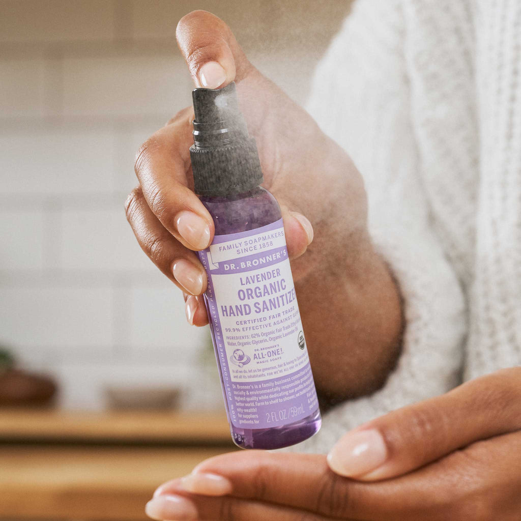 A hand dispenses Dr. Bronners Lavender Organic Hand Sanitizer onto the palm. The scene unfolds in a warmly lit kitchen with tiled walls, emphasizing an eco friendly, vegan, and sustainable lifestyle