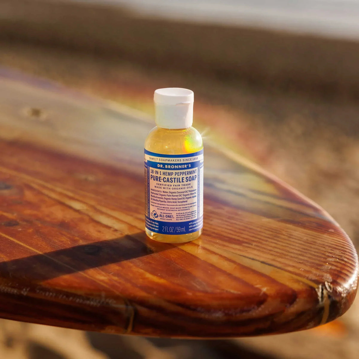 A small bottle of Dr. Bronners 18 in 1 Hemp Peppermint Pure Castile Soap sits on a wooden surfboard in a sunny, beach setting. Dr. Bronners 18 in 1 Hemp Peppermint Pure Castile Soap [2 oz]