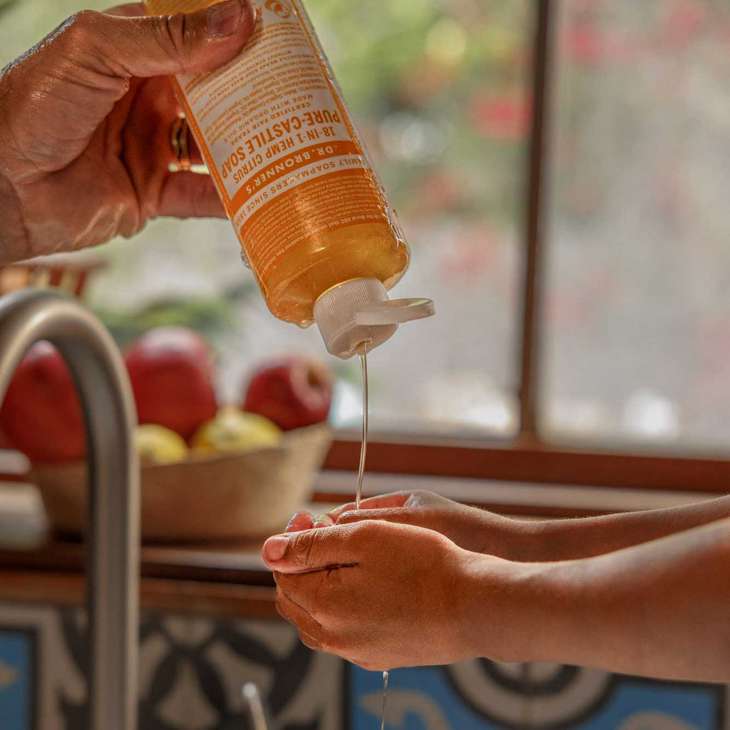 A bottle of Dr. Bronners Pure Castile Soap dispenses liquid onto hands over a kitchen sink. In the background, a window reveals a sunlit garden, and apples are in a bowl [16 oz]