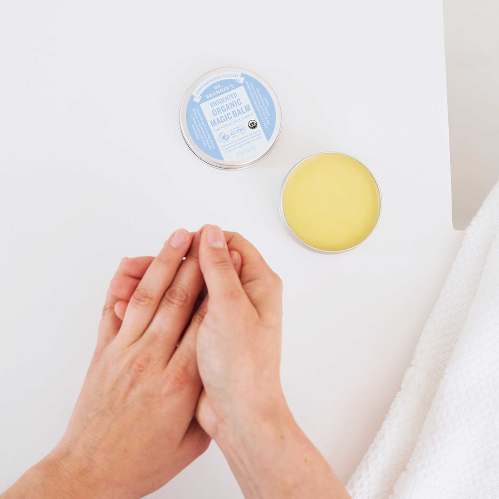 Hands apply balm from an open tin of Dr. Bronners Organic Magic Balm with a blue label on a white surface, next to a textured cloth  [2 oz]