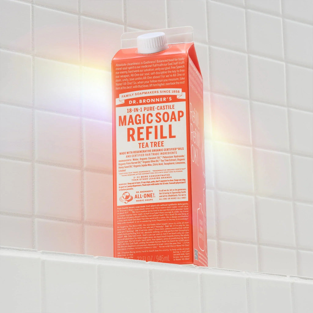 Dr. Bronners 18 in 1 Pure Castile Magic Soap Refill in Tea Tree scent sits upright on a white tiled shelf. The packaging highlights eco friendly and fair trade qualities, emphasizing sustainable practices [32 oz]