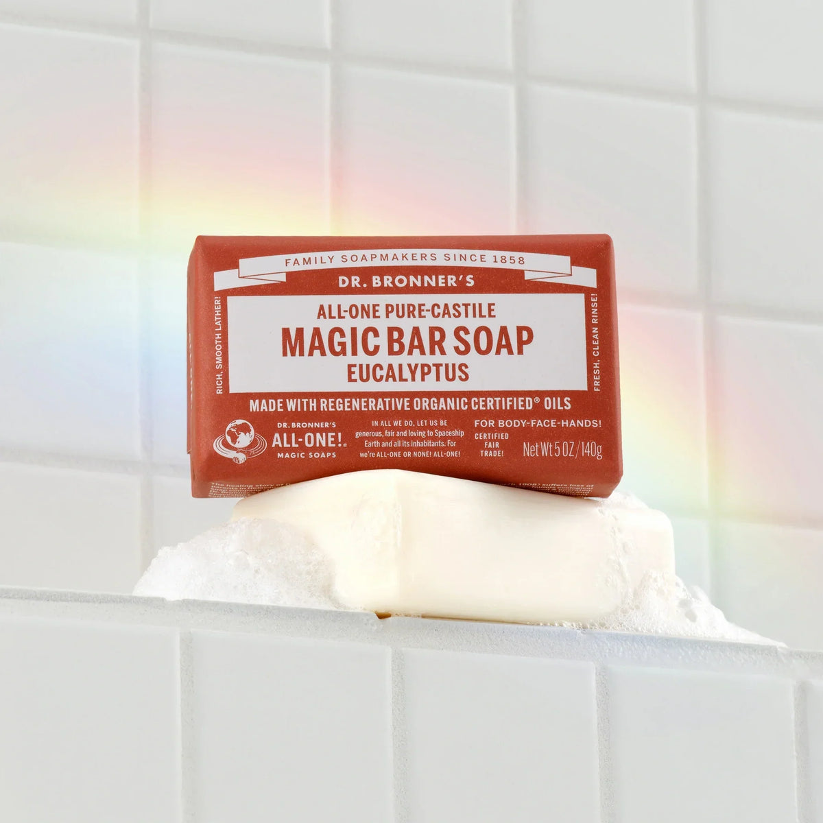 Bar of Dr. Bronners Eucalyptus Magic Bar Soap rests on white tiles with suds and a subtle rainbow effect. Dr. Bronners Magic Bar Soap Eucalyptus, Certified Organic, Fair Trade, Net Wt 5 oz140g [5 oz]
