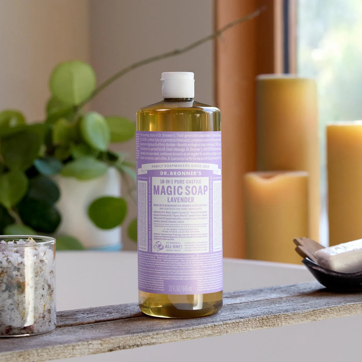 A bottle of Dr. Bronners 18 in 1 Pure Castile Magic Soap in lavender scent stands on a rustic board in a cozy room with candles and greenery, featuring 32 fl oz capacity [32 oz]