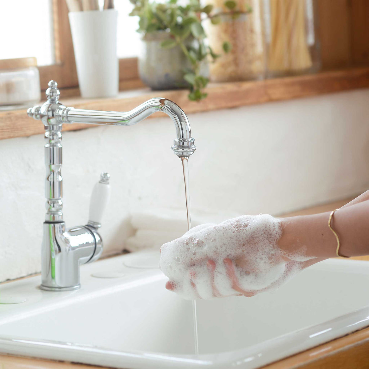 A hand lathers foam near a stainless steel faucet dispensing water into a kitchen sink potted plants and wooden accents subtly enhance the natural, eco friendly ambiance, evoking sustainable soap use [12 oz, 24 oz, 1/2 Gallon]