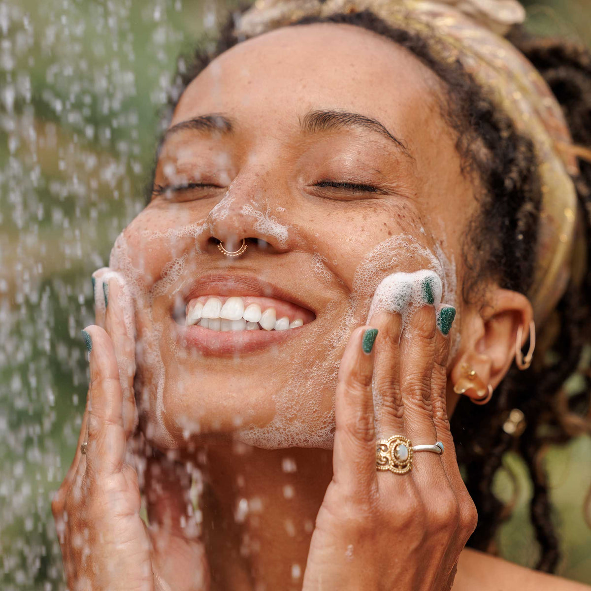 A person gently washes their face with foamy soap, smiling under a shower. The setting is outdoors, with greenery in the background, highlighting a refreshing experience with natural castile soap [32 oz]