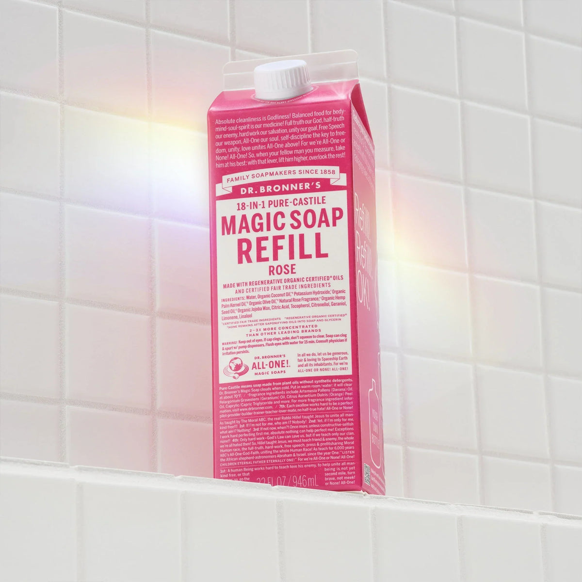 Carton of Dr. Bronners 18 in 1 Pure Castile Magic Soap Refill sits on a white tiled shelf, illuminated by soft sunlight. Dr. Bronners 18 in 1 Pure Castile Magic Soap Refill Rose [32 oz]