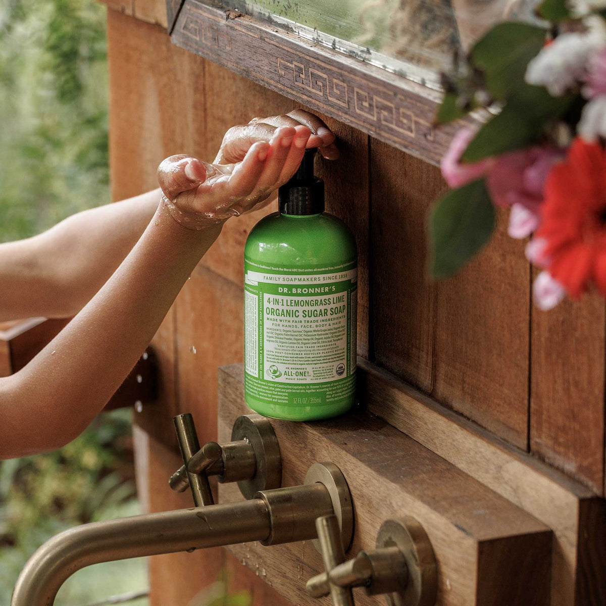 A hand pumps Dr. Bronners 4 in 1 Lemongrass Lime Organic Sugar Soap from a green bottle near a rustic wooden sink with brass fixtures, surrounded by lush greenery and flowers [12 oz]