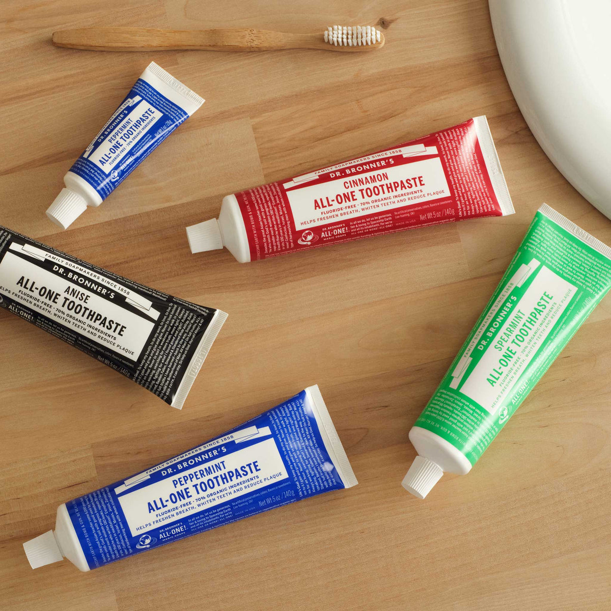 Five tubes of Dr. Bronners toothpaste in various colors lie scattered on a wooden bathroom counter, next to a toothbrush and partially visible sink, suggesting a morning routine setting [5 oz]