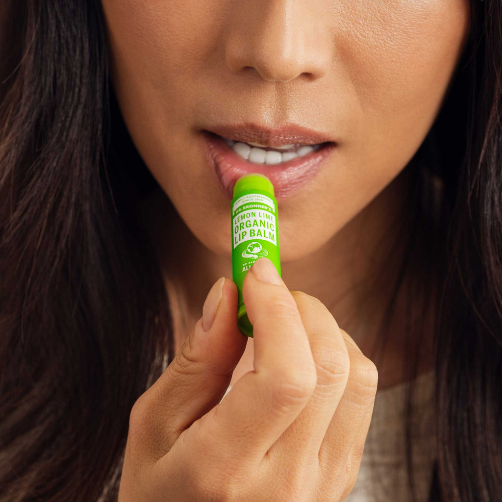 A person applies Dr. Bronners Organic Lip Balm to their lips, holding a green tube labeled Dr. Bronners Magic Organic Lip Balm Lemon Lime. Close up view emphasizes the lip care action [.15 oz]