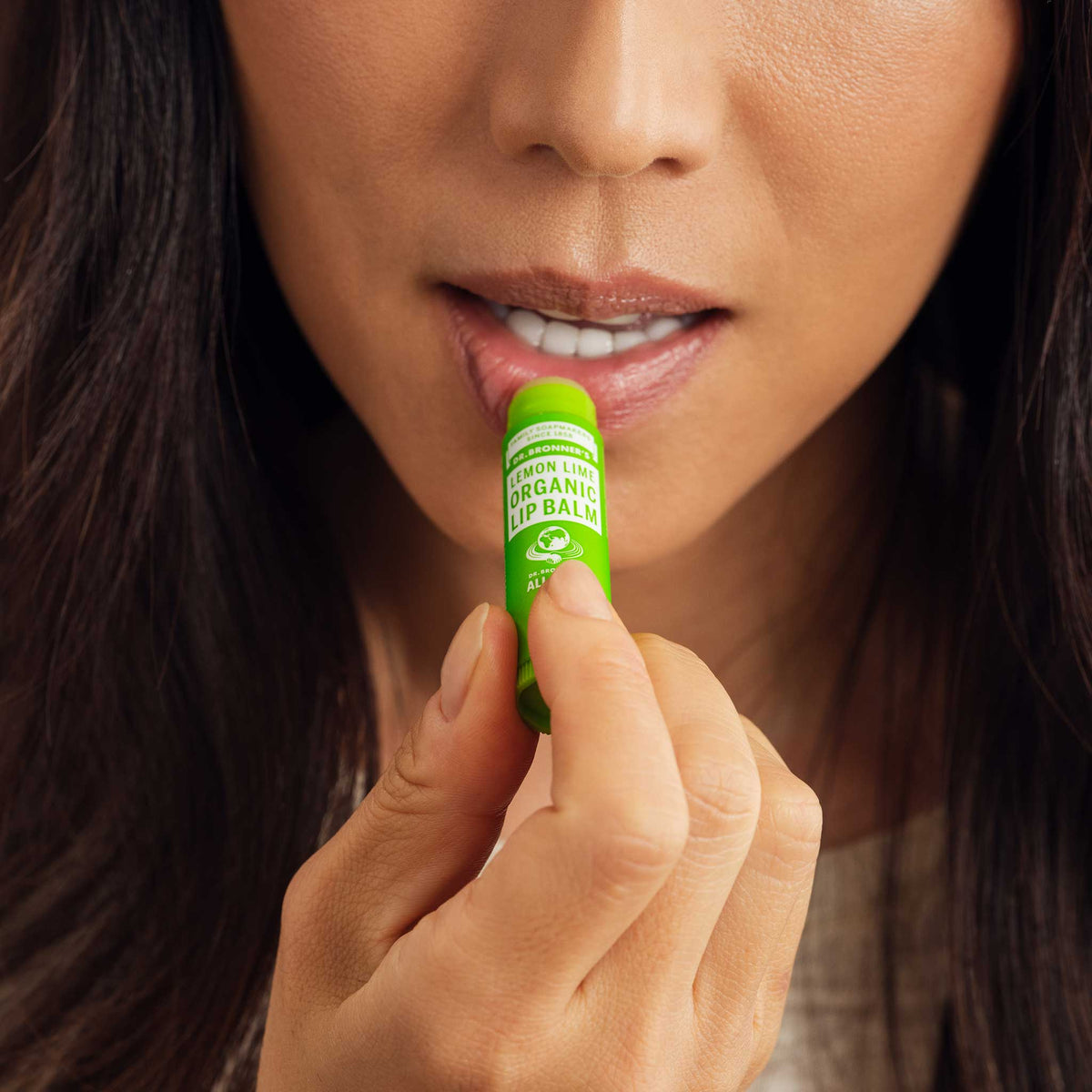 A person applies Dr. Bronners Organic Lip Balm to their lips, holding a green tube labeled Dr. Bronners Magic Organic Lip Balm Lemon Lime. Close up view emphasizes the lip care action [.15 oz]