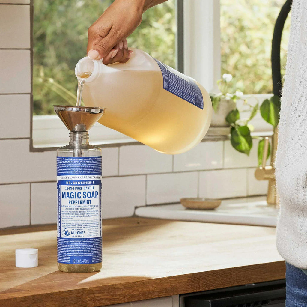 A hand pours Dr. Bronners Pure Castile Soap from a gallon jug into a labeled dispenser in a bright kitchen. Natural light filters through nearby plants, enhancing the clean, eco friendly setting [16 oz]