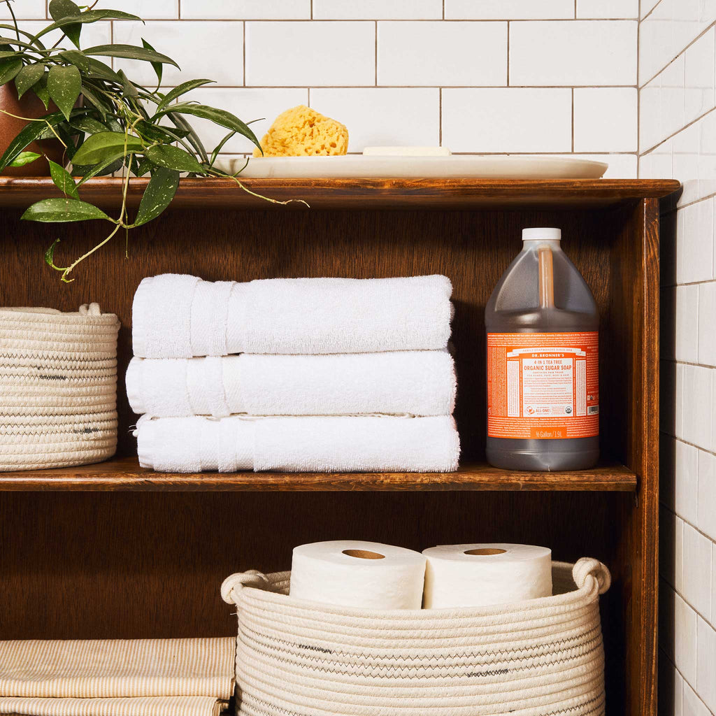 A wooden bathroom shelf displays a large bottle of Dr. Bronners organic sugar soap, white towels, and toilet paper, surrounded by a plant and tile walls, creating a tidy, natural setting [1/2 Gallon]
