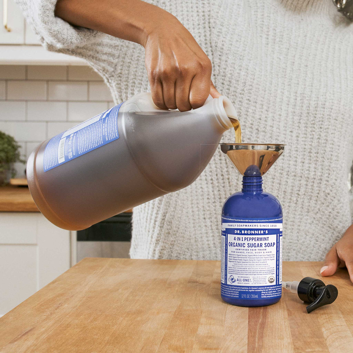 A person pours Dr. Bronners 4 in 1 Peppermint Organic Sugar Soap from a large container into a blue bottle using a funnel on a kitchen counter, surrounded by white tiles and plants [12 oz]