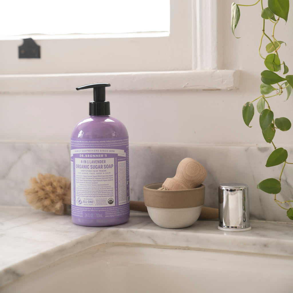 A bottle of Dr. Bronners Lavender Liquid Castile Soap sits on a bathroom countertop alongside a scrubbing brush and potted plant, illuminated by natural light from a nearby window [24 oz]