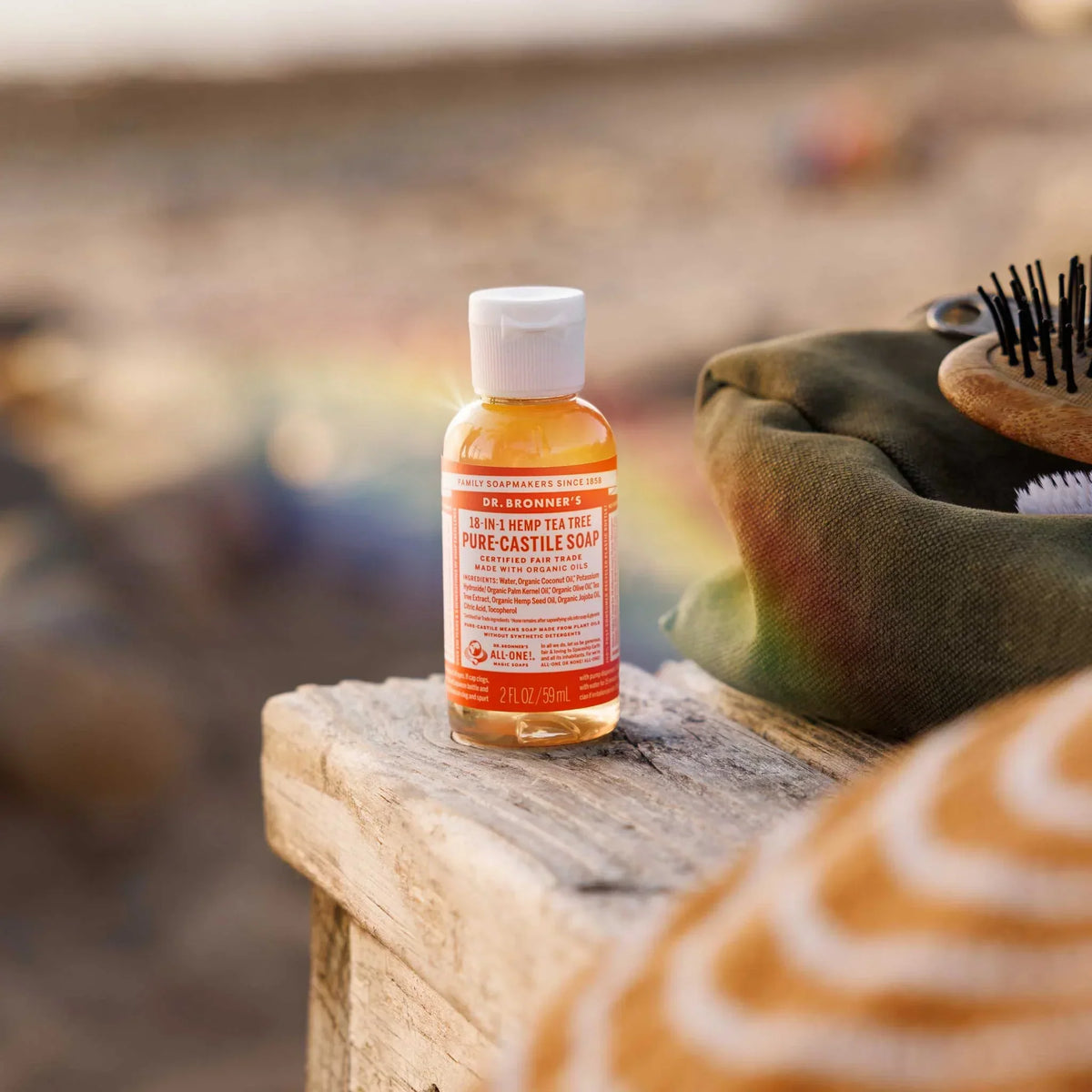 A small bottle of Dr. Bronners 18 in 1 Hemp Tea Tree Pure Castile Soap sits on a wooden surface beside a green bag and a brush, against a blurred outdoor background [2 oz]