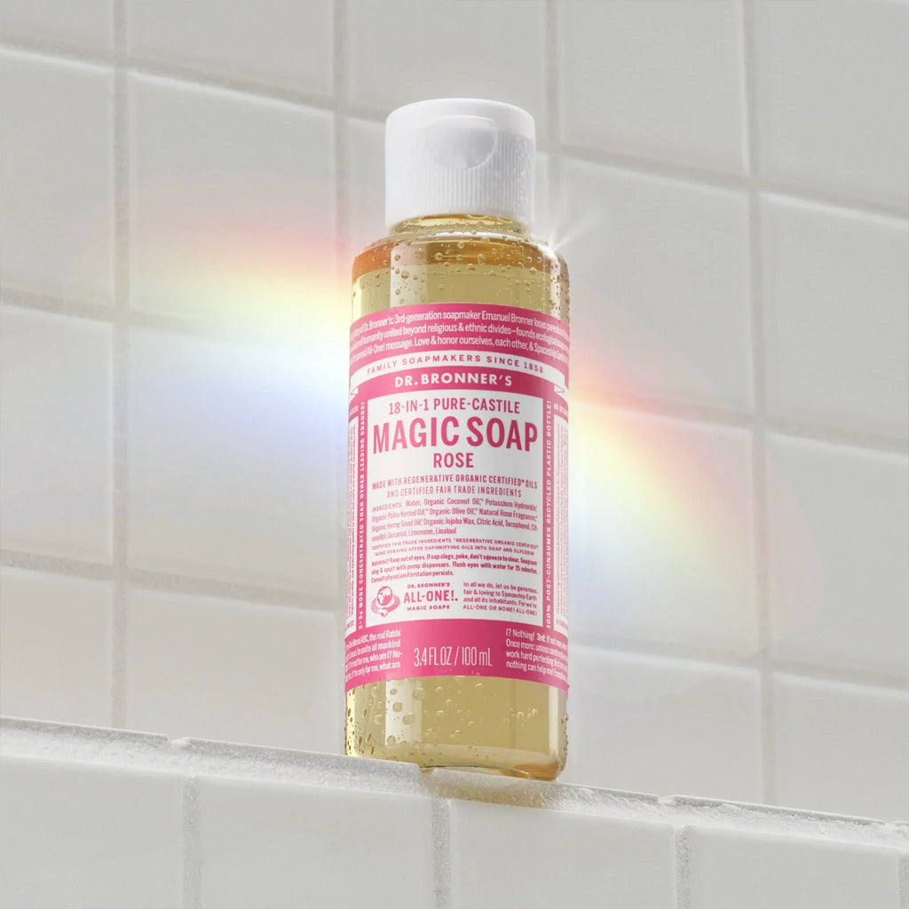 A bottle of Dr. Bronners 18 in 1 Pure Castile Magic Soap, Rose, rests on a white tiled surface. A subtle rainbow effect is visible on the bottle. Text includes 18 IN 1 PURE CASTILE MAGIC SOAP ROSE [3.4 oz]