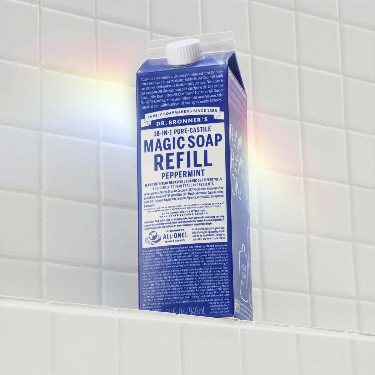 A carton of Dr. Bronners 18 in 1 Pure Castile Magic Soap Refill in peppermint rests on a white tiled surface. Rainbow light reflects softly across the scene. Text includes details about sustainability and use [32 oz]