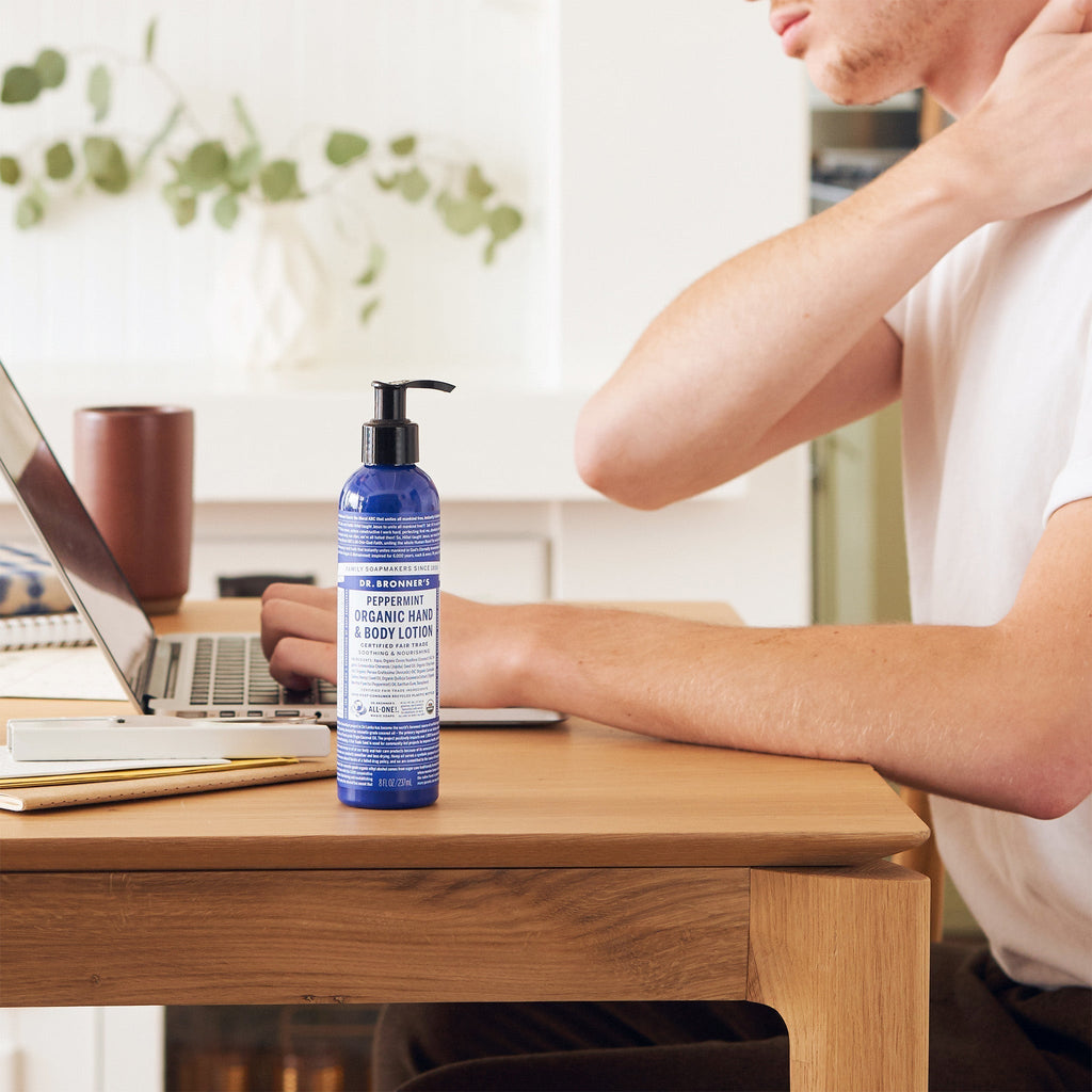 A bottle of Dr. Bronners peppermint organic hand and body lotion sits on a wooden desk next to a person using a laptop, with plants in the background [8 oz]