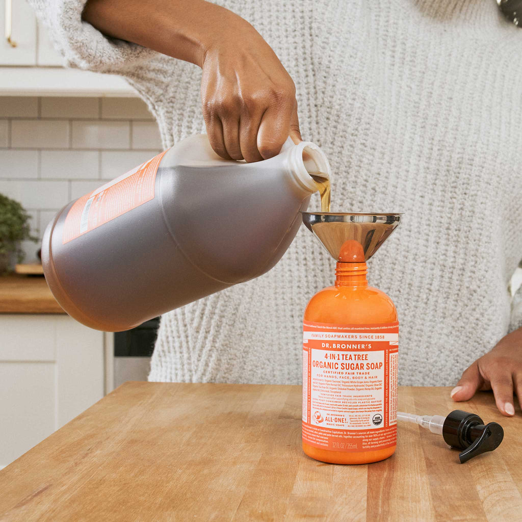 A person pours Dr. Bronners Tea Tree Organic Sugar Soap from a large container into a smaller bottle using a funnel in a bright kitchen. DR. BRONNERS ORGANIC SUGAR SOAP  [12 oz]