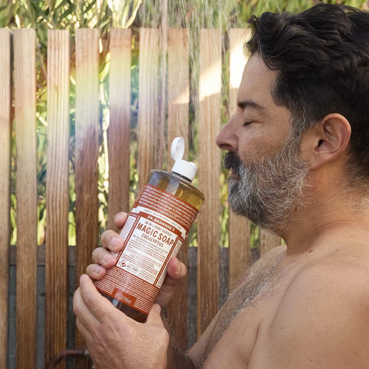 A person holds a bottle of Dr. Bronners Eucalyptus 18 in 1 Pure Castile Soap in a wooden outdoor shower, enjoying the refreshing scent. Text on bottle MAGIC SOAP [32 oz]