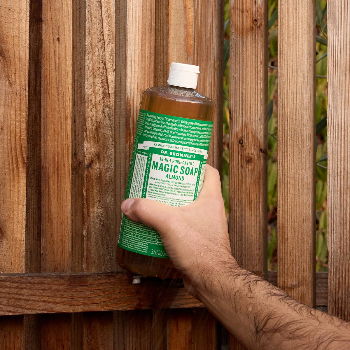 A hand holds a bottle of Dr. Bronners liquid castile soap against a wooden fence. The green label suggests it might be the eucalyptus castile soap. The setting is outdoors [32 oz]