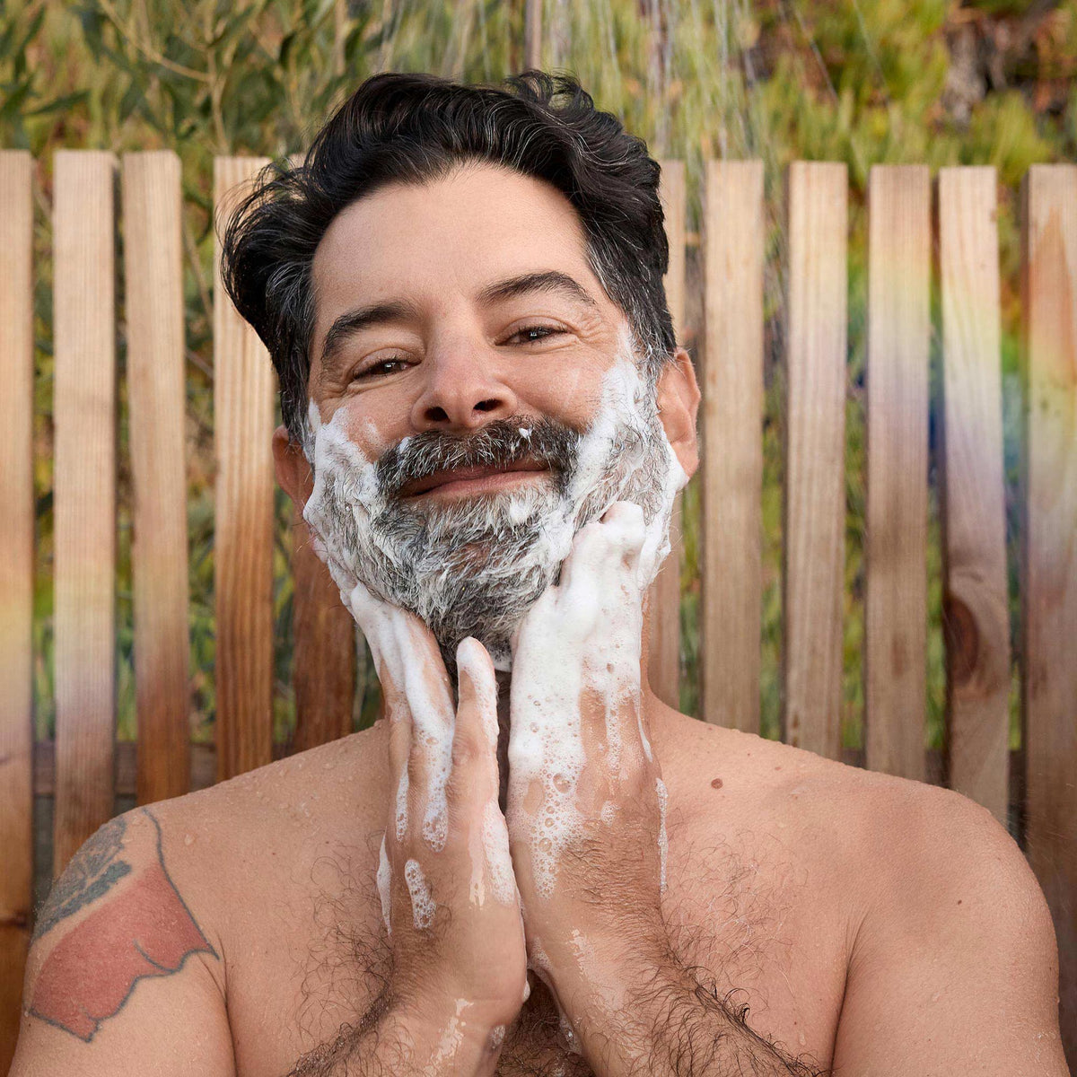 A man lathers his beard with soap while smiling against a wooden fence backdrop, surrounded by greenery. Bathing outdoors suggests a natural and eco friendly lifestyle [32 oz]