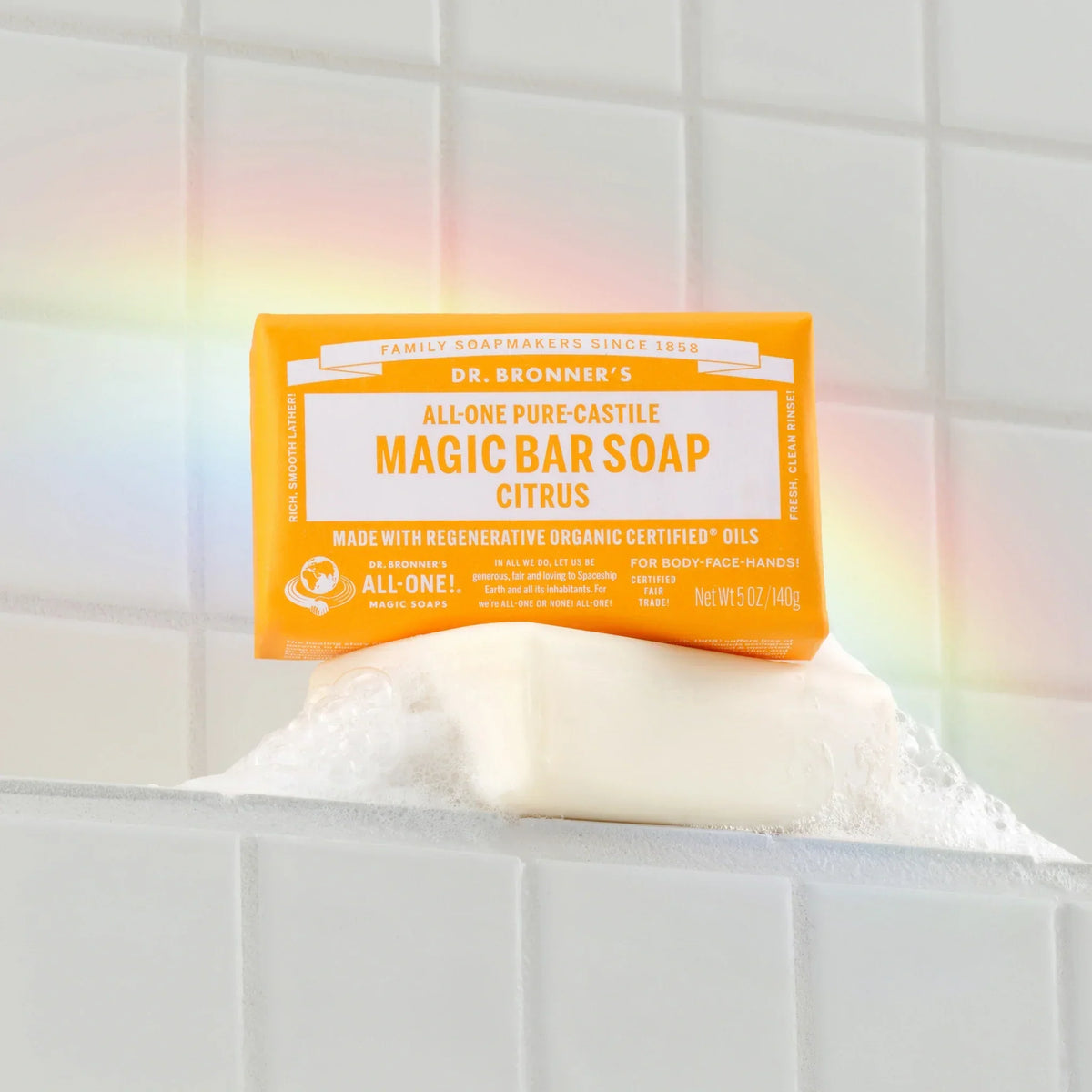 Dr. Bronners citrus magic bar soap rests on a foamy white tile surface with a subtle rainbow glow. All One Pure Castile Magic Bar Soap, Citrus, Made with Regenerative Organic Certified Oils, Net Wt 5 oz140 g [5 oz]