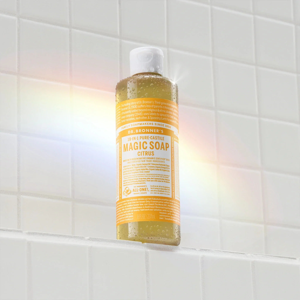 A yellow bottle of Dr. Bronners citrus castile soap sits against a tiled wall, illuminated by a rainbow light reflection. The label features white text detailing the soaps natural ingredients and eco friendly qualities [8 oz]